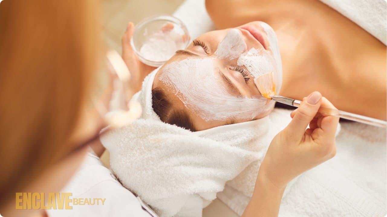 A client relaxing under a white towel while an esthetician brushes a cream mask onto her face
